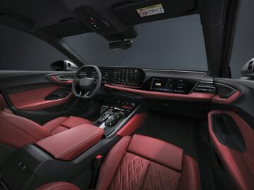 Interior del elegante Audi A5 con asientos de cuero negro y rojo, un salpicadero digital con múltiples pantallas y varios paneles de control. El volante exhibe con orgullo el icónico logotipo de Audi.