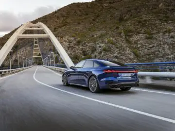 A sleek blue Audi A5 sails across a modern arched bridge surrounded by hilly terrain.