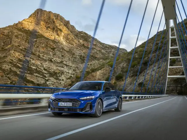 Een Audi A5 rijdt over een moderne brug met kabels, tegen een achtergrond van rotsachtige bergen.