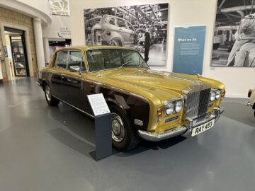 Een gouden en crèmekleurige vintage Rolls-Royce wordt binnenshuis tentoongesteld met informatieborden in het British Motor Museum. De klassieke auto, met zijn prominente grille, staat trots op een grijze vloer.