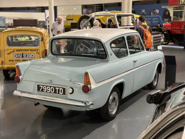 Een vintage lichtblauwe Ford Anglia-auto met kenteken "7990 TD" wordt tentoongesteld in de showroom van het British Motor Museum, omringd door andere voertuigen en verschillende mensen.