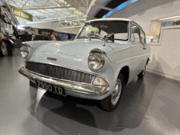 Een lichtblauwe vintage auto met een Brits kenteken "7990 TD" is te zien in een overdekte showroom in het British Motor Museum. De auto heeft ronde koplampen en een chromen grille, waardoor het een hoogtepunt is voor wie klassiek autodesign bezoekt en waardeert.