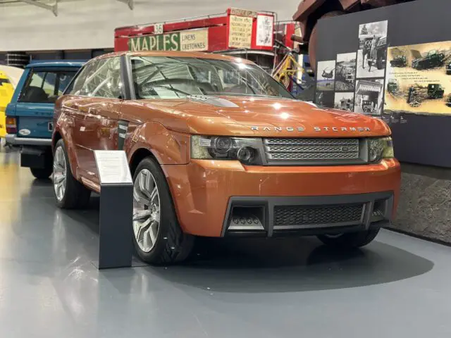 Een oranje Range Stormer-conceptauto is te zien in het British Motor Museum. Informatieborden geven details terwijl andere voertuigen op de achtergrond deze fascinerende autoreportage voor bezoekers vervolledigen.