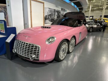 Een grote roze auto in vintagestijl met zes wielen staat tentoongesteld in het British Motor Museum. De voorkant van de auto is voorzien van een opvallende grille en ronde koplampen. In deze reportage zie je op de achtergrond andere klassieke auto's zichtbaar, die elk hun eigen verhaal uit de autogeschiedenis vertellen.