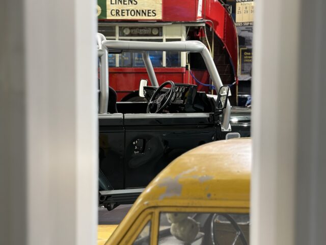 Een gele auto met een versleten exterieur op de voorgrond, een blauwe terreinwagen en een rode dubbeldekkerbus met reclame voor "Linens Cretonnes" op de achtergrond, bekeken door een smalle opening tijdens ons reportagebezoek aan het British Motor Museum.