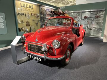 Een felrode vintage auto met het kenteken "TFC 963" wordt tentoongesteld in een museumtentoonstelling, te midden van verschillende auto-artefacten, in het British Motor Museum.