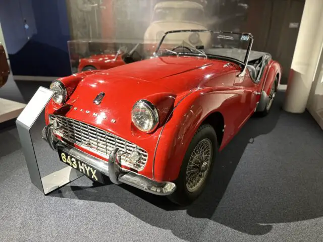 Een klassieke rode cabriolet, geïdentificeerd als model 643 HXX, wordt tentoongesteld in het British Motor Museum op een vloer met vloerbedekking en biedt bezoekers een nostalgische ervaring.