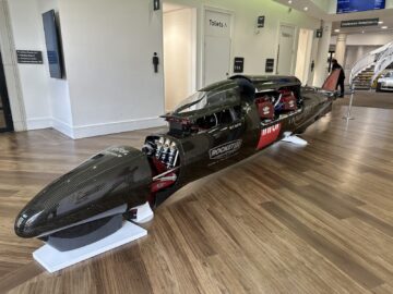 Een strakke, zwarte raketauto die binnen in het British Motor Museum te zien is, geplaatst op een houten vloer nabij de toiletten en een conferentiereceptieruimte, waardoor het een intrigerend hoogtepunt is voor bezoekers.