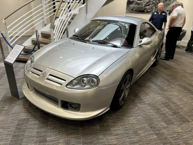 Een zilveren sportwagen wordt tentoongesteld in een museumtentoonstelling in het British Motor Museum, met op de achtergrond twee mensen in gesprek bij een trap.