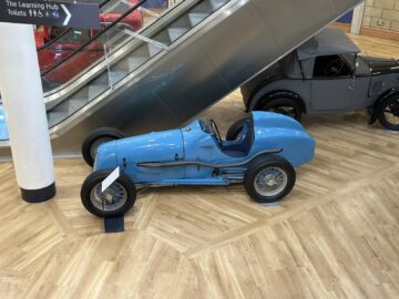 In een tentoonstelling met houten vloeren nabij de roltrap staan een vintage blauwe raceauto en een grijze klassieke auto trots tentoongesteld, die doet denken aan de strakke elegantie van het British Motor Museum.