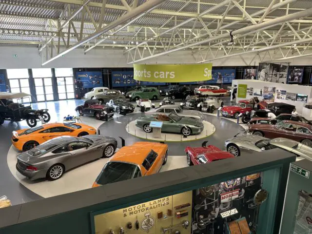 Een ruime showroom in het British Motor Museum biedt een verscheidenheid aan klassieke en moderne auto's, waaronder sportwagens, in een goed verlicht gebouw met hoge plafonds en een groen bord met de tekst 'sportwagens' erboven.