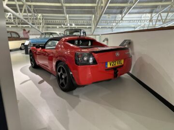 Een rode sportwagen met een kentekenplaat met de tekst "V22 VXL" staat binnen geparkeerd nabij een witte muur bij het British Motor Museum. Op de achtergrond zijn andere klassieke auto's zichtbaar, waardoor dit pittoreske tafereel een bezoek waard is.