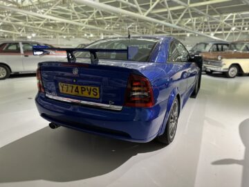 Een blauwe sportwagen met spoiler staat binnen geparkeerd bij het British Motor Museum. Op het kenteken staat 'Y774 PVS'. Op de achtergrond zijn andere klassieke auto's zichtbaar, waardoor een boeiend tafereel ontstaat dat perfect is voor een reportage over de autogeschiedenis.