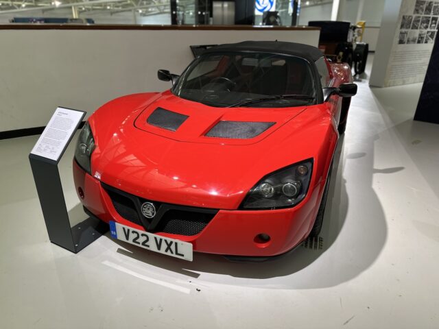 Een rode sportwagen met kenteken "V22 VXL" wordt binnenshuis tentoongesteld in het British Motor Museum, vergezeld van een informatiestand ernaast. De achtergrond omvat een deel van een tentoonstellingsruimte met informatiepanelen, die bezoekers uitnodigen om in een fascinerend bezoek te duiken.