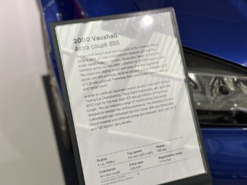 Een displaykaart van de Vauxhall Astra Coupé 888 uit 2000 in het British Motor Museum, met gedetailleerde technische specificaties en kenmerken, met een deel van de blauwe auto op de achtergrond.