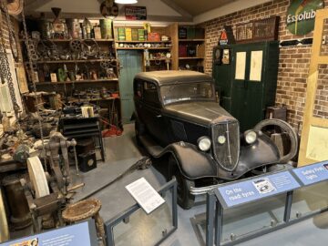 Een vintage zwarte auto wordt tentoongesteld in een museumwerkplaats in het British Motor Museum, omringd door verschillende antieke gereedschappen en uitrusting op planken en muren. Informatieborden worden voor het voertuig geplaatst en bieden bezoekers een meeslepende bezoekervaring.