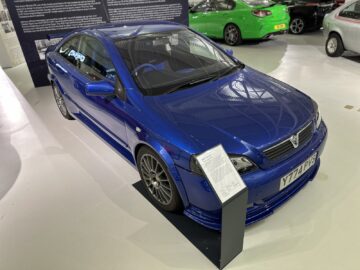 Een blauwe sportwagen, tentoongesteld in een overdekte tentoonstellingsruimte van het British Motor Museum, springt in het oog; Voor de auto wordt een informatiebord geplaatst, met details over de rijke geschiedenis en specificaties die alle bezoekers kunnen waarderen.