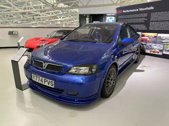 Een blauwe Vauxhall-prestatieauto met kenteken Y774 PVS is te zien in een showroom in het British Motor Museum. Op de achtergrond is een rode auto zichtbaar, naast een displaypaneel met de titel 'Performance Vauxhalls', dat perfect het thema van deze opwindende reportage weergeeft.