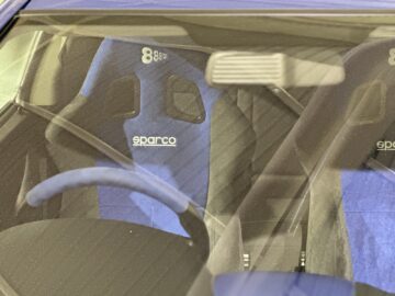 Close-up van het interieur van een auto met Sparco-racestoelen met blauwe bekleding, zichtbaar stuur en een deel van de achteruitkijkspiegel door de voorruit, die doet denken aan een klassiek display dat je in het British Motor Museum zou vinden.