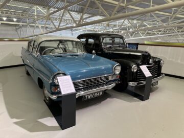 Twee klassieke auto's, een blauw-witte sedan en een zwarte sedan, worden binnenshuis tentoongesteld met informatieborden ervoor. Tijdens ons bezoek aan het British Motor Museum ontdekten we dat deze meesterwerken de essentie van de autogeschiedenis weergeven.