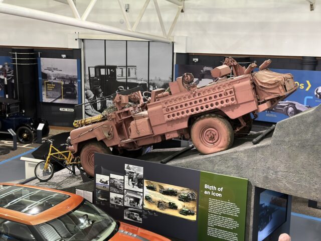 Een roze, gepantserde jeep staat op een helling tentoongesteld in een museumtentoonstelling in het British Motor Museum. In de buurt staan informatiepanelen en andere vintage voertuigen die bezoekers kunnen verkennen.