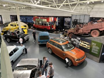 Een verscheidenheid aan vintage en moderne voertuigen is te zien in een grote overdekte tentoonstellingsruimte in het British Motor Museum, waaronder auto's, vrachtwagens en een militaire tank. Verschillende mensen observeren tijdens hun bezoek de tentoongestelde voorwerpen.