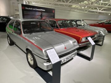 Een rij klassieke Vauxhall-auto's tentoongesteld in een showroom in het British Motor Museum met beschrijvende platen voor elk voertuig. De dichtstbijzijnde auto is een zilveren hatchback met een rode streep.