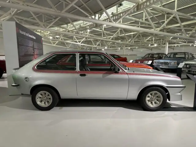 Tijdens ons bezoek aan het British Motor Museum bewonderden we een klassieke zilveren hatchback met zwarte en rode strepen, elegant tentoongesteld in een museumsetting en omringd door andere vintage voertuigen.