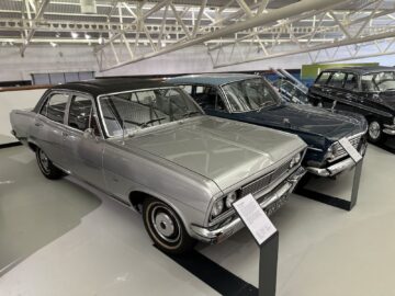 Twee klassieke sedans worden tentoongesteld in een overdekte autotentoonstelling in het British Motor Museum. De voorste auto is zilverkleurig en de achterste donkerblauw, elk met informatieve borden ernaast, die bezoekers een gedetailleerde reportage van hun geschiedenis bieden.