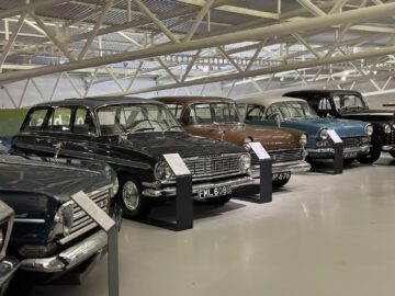 Binnen in het British Motor Museum staat een rij oldtimers tentoongesteld, elk op een verhoogd platform met informatieve borden ervoor. De auto's variëren in kleuren, waaronder zwart, bruin en blauw. Tijdens uw bezoek kunt u fascinerende details leren over de geschiedenis van elk voertuig.