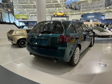 Tijdens ons bezoek aan het British Motor Museum verwonderden we ons over een groene hatchback die in een overdekte showroom stond naast andere oldtimers, waaronder een beige compacte auto en een witte coupé, allemaal onder een elegant hoge plafondconstructie.