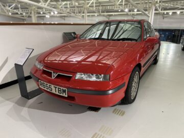 Een rode klassieke auto met een kentekenplaat met de tekst "R655 TBM" wordt binnenshuis tentoongesteld in het British Motor Museum. Naast de auto zijn informatiestands geplaatst.