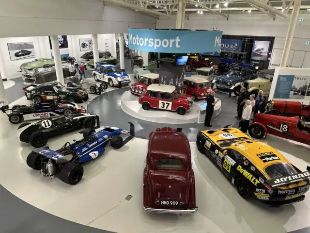 Een motorsporttentoonstelling in het British Motor Museum toont verschillende raceauto's, waaronder klassieke en moderne ontwerpen. Verschillende bezoekers bekijken en fotograferen de auto's, terwijl prominent een groot bord 'Motorsport' zichtbaar is.