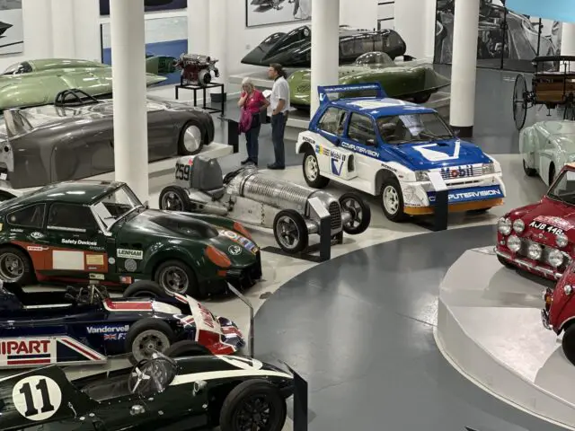 In de tentoonstelling van het British Motor Museum zijn verschillende vintage- en raceauto's te zien. Er worden twee mensen gezien die de voertuigen observeren, waaronder een opvallende blauw-witte rallyauto op de voorgrond.