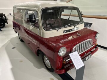 Een vintage Bedford CA Van, tentoongesteld in het British Motor Museum, heeft een tweekleurig crème- en kastanjebruin kleurenschema, met een informatiebord ervoor.