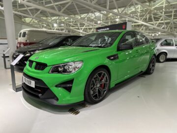 Een felgroene sedan met zwarte details wordt binnenshuis tentoongesteld in het British Motor Museum. De auto heeft een strak ontwerp, rode remklauwen en een "VRX" kentekenplaat. Op de achtergrond zijn andere voertuigen zichtbaar, wat de allure van deze autoreportage vergroot.