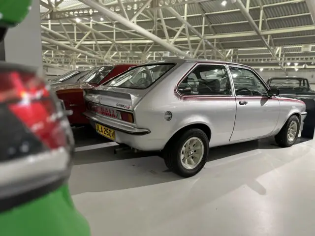 Een zilveren vintage auto met kenteken JAJ 250V staat binnen geparkeerd in een goed verlichte faciliteit tussen andere klassieke voertuigen, waardoor dit het hoogtepunt is van elk bezoek aan het British Motor Museum.