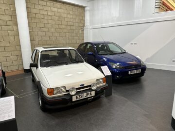 In een overdekte showroom van het British Motor Museum staan twee oldtimers tentoongesteld: een witte Ford met kenteken E131 JPA aan de linkerkant en een blauwe Ford met kenteken BN54 GXK aan de rechterkant, waardoor bezoekers een meeslepend en nostalgisch bezoek krijgen.