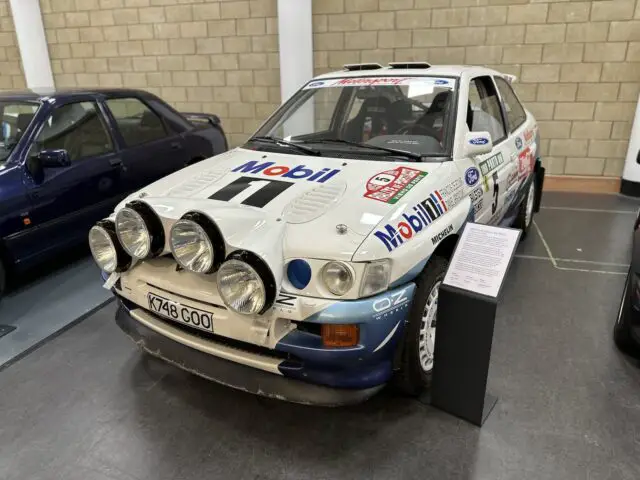 Een witte rallyauto met meerdere schijnwerpers aan de voorkant en verschillende sponsorlogo's op de carrosserie wordt binnenshuis tentoongesteld naast een informatiebord in het British Motor Museum, perfect voor degenen die besluiten een bezoek te brengen en meer te leren over de legendarische geschiedenis ervan.