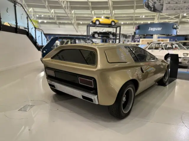 Een futuristische lichtbruine conceptauto met zwarte accenten aan de achterkant wordt tentoongesteld in een overdekte autotentoonstelling in het British Motor Museum, met andere klassieke en moderne auto's zichtbaar op de achtergrond. Bezoekers kunnen genieten van deze opmerkelijke autoshow als onderdeel van hun reis door de uitgebreide collectie van het museum.