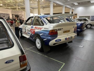 Een Ford-rallyauto met sponsorstickers is te zien in het British Motor Museum. Op de achtergrond zijn andere oldtimers zichtbaar en is een kleine groep bezoekers te zien.