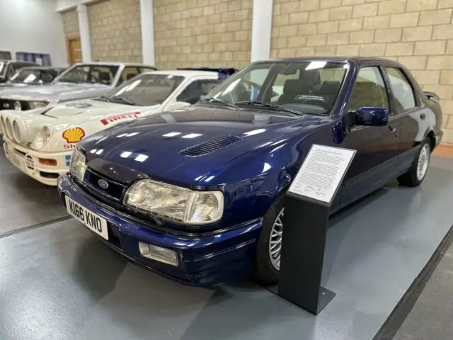 Een donkerblauwe klassieke Ford-auto wordt binnen tentoongesteld naast andere vintage voertuigen in het British Motor Museum. Ernaast staat een bordje met informatie, dat bezoekers een boeiende ervaring biedt tijdens hun bezoek.