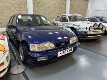 Een blauwe Ford-auto met kenteken "K166 KNO" wordt binnen tentoongesteld naast een witte rallyauto versierd met meerdere koplampen en emblemen, die doet denken aan een scène die je tegenkomt tijdens een bezoek of reportage in het British Motor Museum.