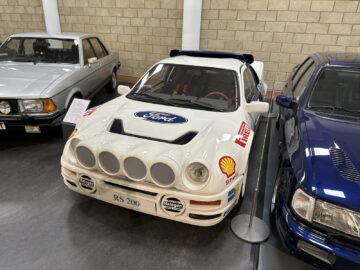 Een witte Ford RS200-rallyauto wordt binnenshuis tentoongesteld naast andere voertuigen in het British Motor Museum, met bakstenen muren op de achtergrond. De grille aan de voorzijde is voorzien van grote mistlampen en sponsorlogo's, wat zorgt voor een spannend bezoek, perfect voor uw volgende bezoek of reportage.