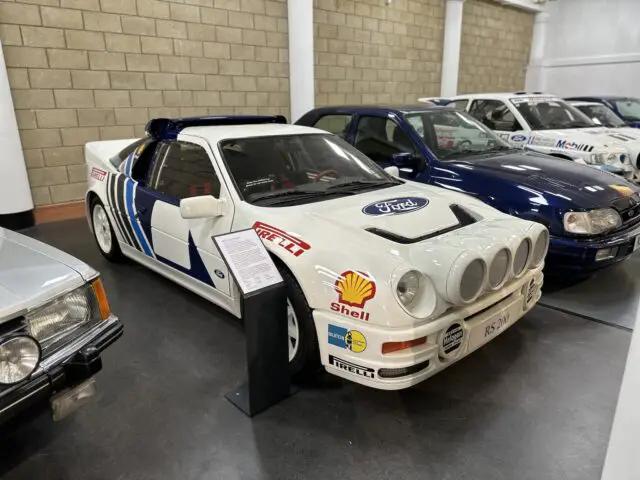 Een witte Ford RS200-rallyauto met blauwe emblemen wordt binnen tentoongesteld in het British Motor Museum, naast andere voertuigen. De auto is voorzien van sponsorlogo’s van Shell en Pirelli en is voorzien van opvallende koplampen aan de voorzijde. Bezoekers kunnen genieten van een uitgebreide reportage over de geschiedenis ervan.