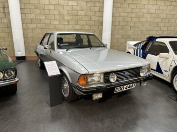 Een zilveren vintage sedan met een informatiekaart staat binnen tegen een bakstenen muur. Op het kenteken staat "EOO 444T." Dit zeldzame juweeltje, perfect voor elk bezoek aan het British Motor Museum, is een stille getuige van de autogeschiedenis.