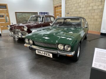 Twee klassieke auto's, een groene Ford Capri en een kastanjebruine auto, worden binnenshuis tentoongesteld op een grijze vloer in het British Motor Museum. Op de voorgrond bij de Ford Capri is een beschrijvingsplaatje zichtbaar, waardoor bezoekers een inzichtelijke reportage over de geschiedenis ervan krijgen.