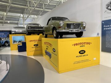 Een reportagewaardige tentoonstelling van oldtimers in het British Motor Museum toont de ‘Prototype Series’ op gele platforms, met een MG Mini ADO 34 uit 1964 prominent op een hoger niveau. Op de achtergrond zijn bordjes en extra auto's te zien, waardoor het een must-see is voor elk bezoek.