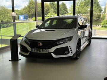 Een witte Honda Civic Type R wordt tentoongesteld op een showroomvloer met grote ramen en groen zichtbaar aan de buitenkant, alsof hij is voorbereid op een bezoek of zelfs een reportage van het British Motor Museum.