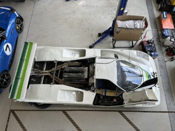 Een bovenaanzicht van een witte raceauto met groene strepen, geparkeerd in een garage bij het British Motor Museum, met de motorkap aan de achterkant gedeeltelijk open. Verschillende gereedschappen en apparatuur zijn verspreid over het gebied en leggen een openhartig moment in reportagestijl vast.